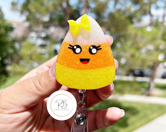 Può includere: Un portabadge a forma di caramella di mais con un volto sorridente, ciglia e un fiocco giallo. Il portabadge è giallo, arancione e bianco con glitter. Il portabadge è tenuto in una mano.