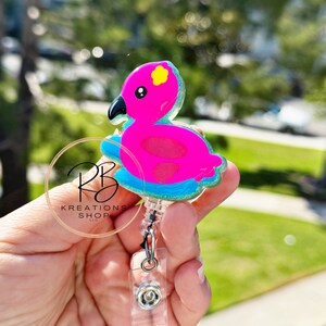 May include: A pink flamingo-shaped badge reel with a black beak and yellow accents. The badge reel has a clear plastic retractable cord and a clear plastic clip. The badge reel is held in a hand.