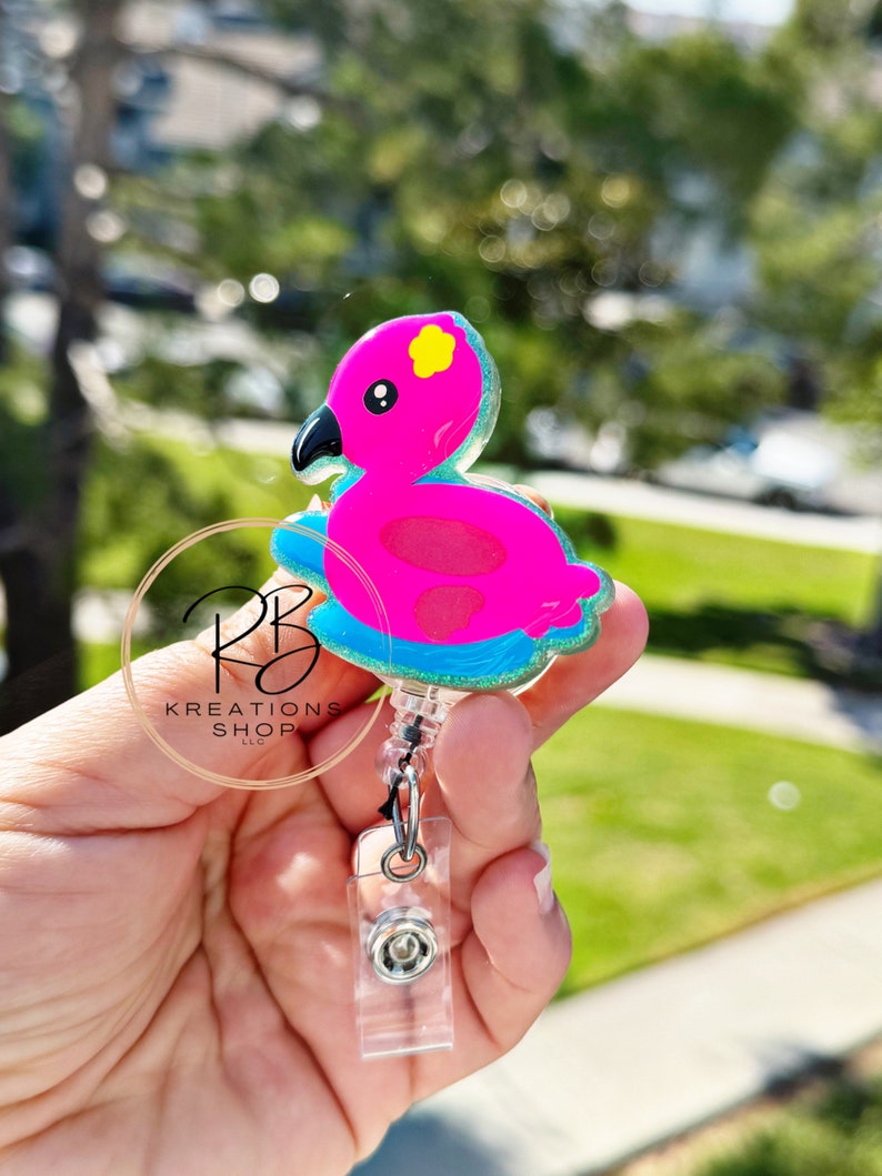 May include: A pink flamingo-shaped badge reel with a black beak and yellow accents. The badge reel has a clear plastic retractable cord and a clear plastic clip. The badge reel is held in a hand.