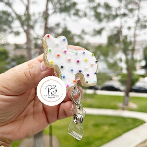 May include: A white, cat-shaped badge reel with colorful dot accents and gold glitter trim. The badge reel has a clear plastic strap and a metal clip. The badge reel is from the PB Kreations Shop.