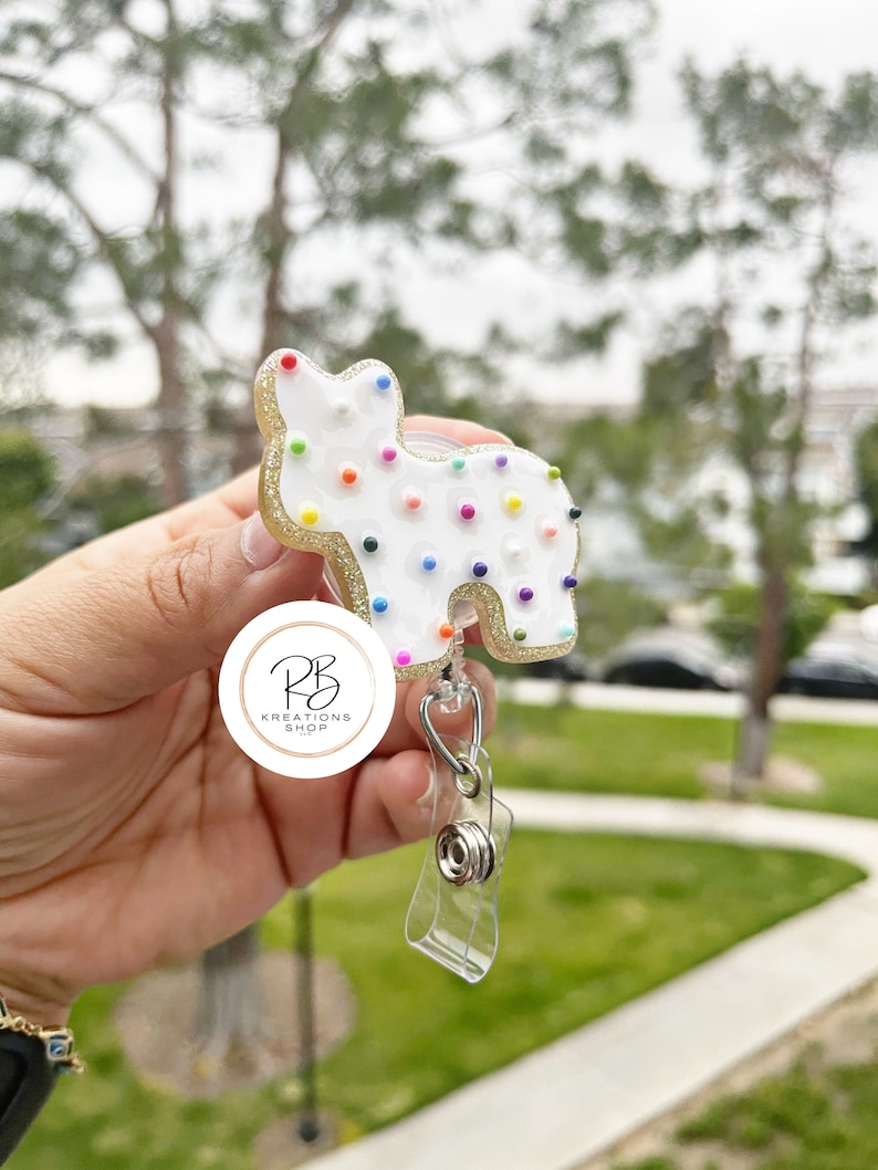 May include: A white, cat-shaped badge reel with colorful dot accents and gold glitter trim. The badge reel has a clear plastic strap and a metal clip. The badge reel is from the PB Kreations Shop.