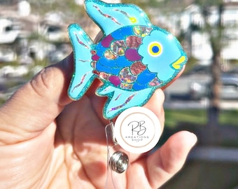 May include: A blue and purple glitter fish charm with a yellow eye, attached to a clear retractable badge holder with a white circle that says "RB Kreations Shop".