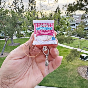May include: A red and white Uncrustables peanut butter and strawberry jam sandwich keychain. The keychain is attached to a clear plastic retractable badge holder.