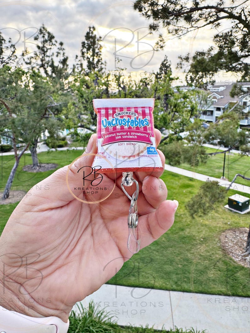 May include: A red and white Uncrustables peanut butter and strawberry jam sandwich keychain. The keychain is attached to a clear plastic retractable badge holder.