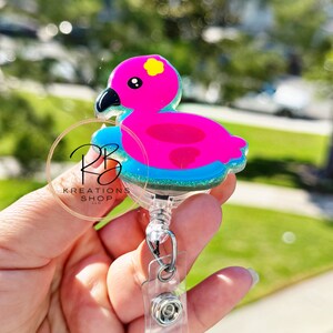 May include: A pink flamingo-shaped badge reel with a black beak and yellow flower detail. The flamingo floats on a blue glittery base. The reel has a clear plastic clip and a retractable cord. The text "RB Kreations Shop" is visible.