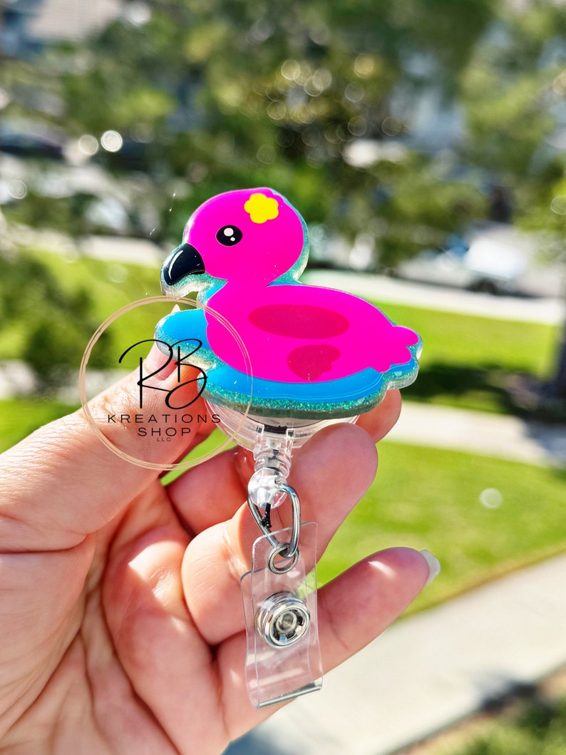 May include: A pink flamingo-shaped badge reel with a black beak and yellow flower detail. The flamingo floats on a blue glittery base. The reel has a clear plastic clip and a retractable cord. The text "RB Kreations Shop" is visible.