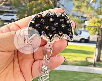 May include: A black bat-shaped badge holder with a gold glitter border and colorful dots. The holder is attached to a clear retractable badge reel.