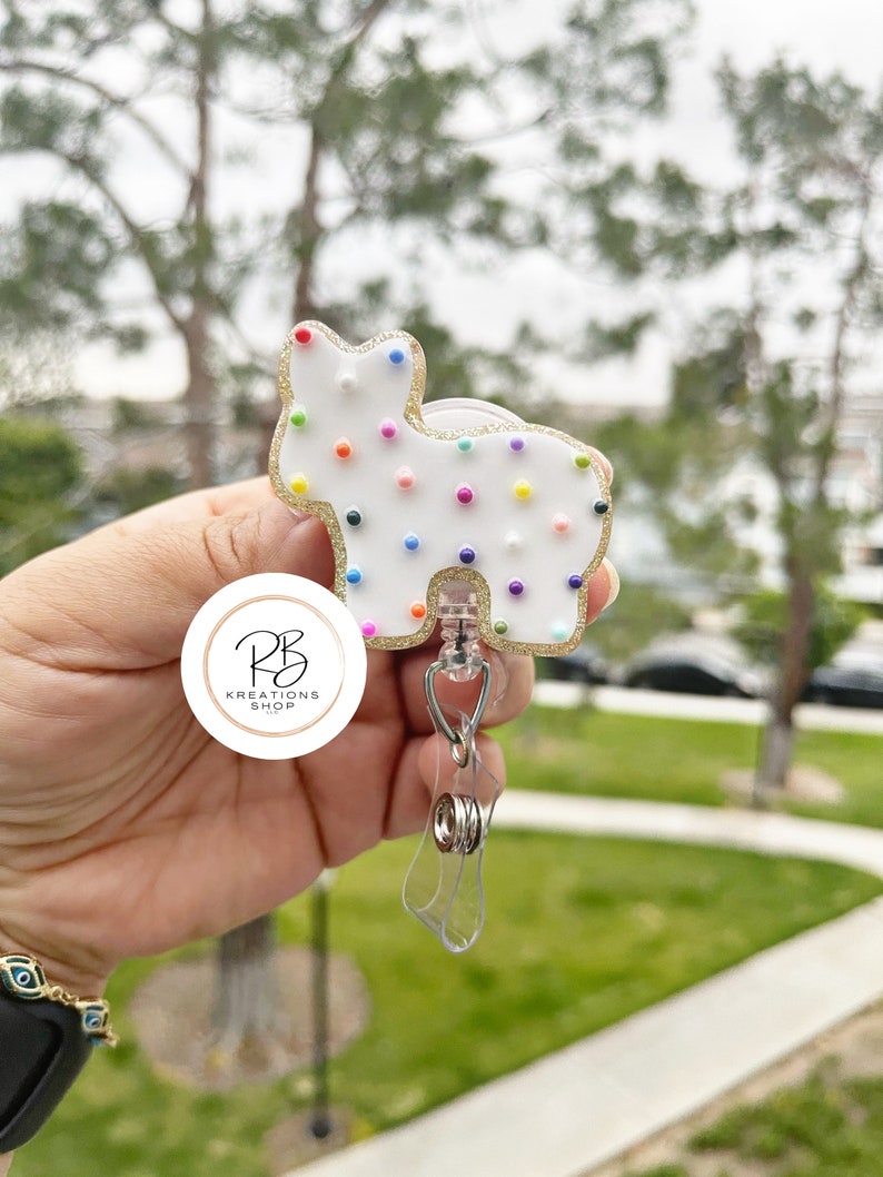 May include: A white, cat-shaped badge reel with colorful dot accents and a gold glitter outline. The reel has a clear plastic retractable cord and a metal clip. The badge reel has the text "RB Kreations Shop" in a circular logo.