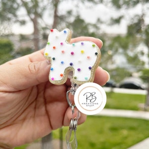 May include: A white and gold badge reel in the shape of a cookie with colorful sprinkles. The badge reel has a retractable cord and a clear plastic clip. The badge reel has the text "RB Kreations Shop" in a circular logo.