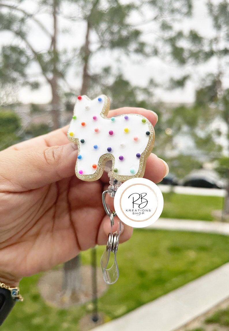May include: A white and gold badge reel in the shape of a cookie with colorful sprinkles. The badge reel has a retractable cord and a clear plastic clip. The badge reel has the text "RB Kreations Shop" in a circular logo.
