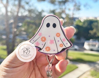 May include: A white ghost-shaped badge holder with pink and orange floral accents and glitter. The holder is attached to a clear retractable badge reel.