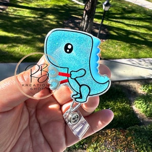 May include: A light blue glitter dinosaur with a red ribbon around its neck. The dinosaur is attached to a clear plastic retractable badge holder.