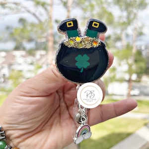 May include: A black pot of gold with a green shamrock and two green leprechaun hats with gold buckles. The pot of gold is attached to a silver retractable badge holder with a rainbow charm. The badge holder has a white circle with the text "PB KREATIONS SHOP".