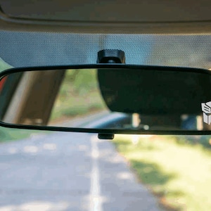 May include: A rearview mirror with a white Transformers logo sticker on the right side. The mirror is reflecting a road and trees.