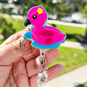 May include: A pink flamingo badge reel with a yellow flower accent on its head. The flamingo is on a blue floatie with a glittery border. The badge reel has a clear plastic clip and a retractable cord.