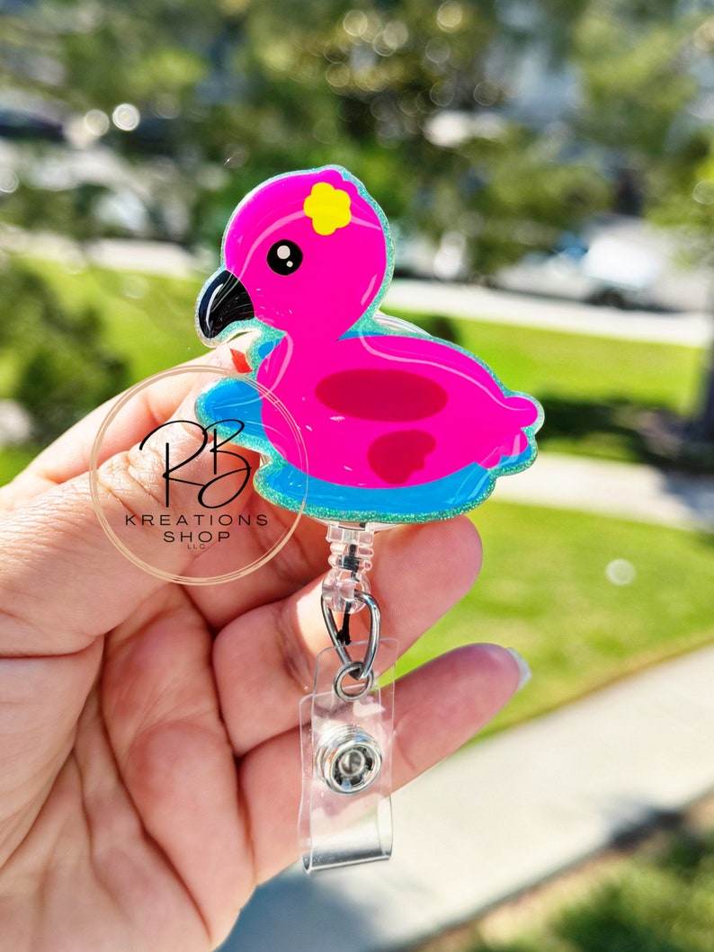 May include: A pink flamingo badge reel with a yellow flower accent on its head. The flamingo is on a blue floatie with a glittery border. The badge reel has a clear plastic clip and a retractable cord.