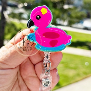 May include: A pink flamingo badge reel with a yellow flower accent and black beak. The badge reel has a clear plastic clip and a retractable cord. The text "RB Kreations Shop LLC" is visible on a clear circular tag.