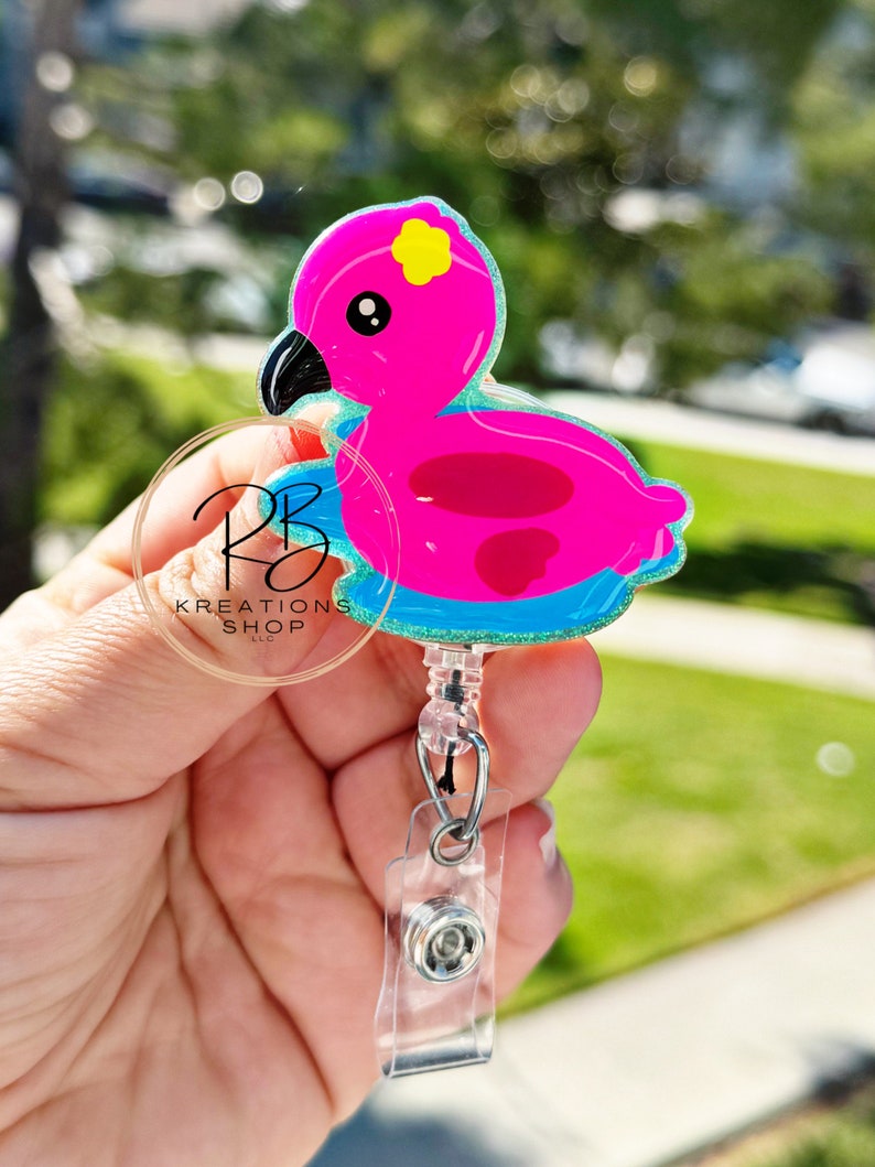 May include: A pink flamingo badge reel with a yellow flower accent and black beak. The badge reel has a clear plastic clip and a retractable cord. The text "RB Kreations Shop LLC" is visible on a clear circular tag.