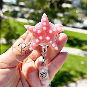 May include: A pink starfish-shaped badge reel with white polka dots. The badge reel has a clear plastic retractable cord and a metal clip. The logo "RB Kreations Shop LLC" is printed on a clear plastic disc.