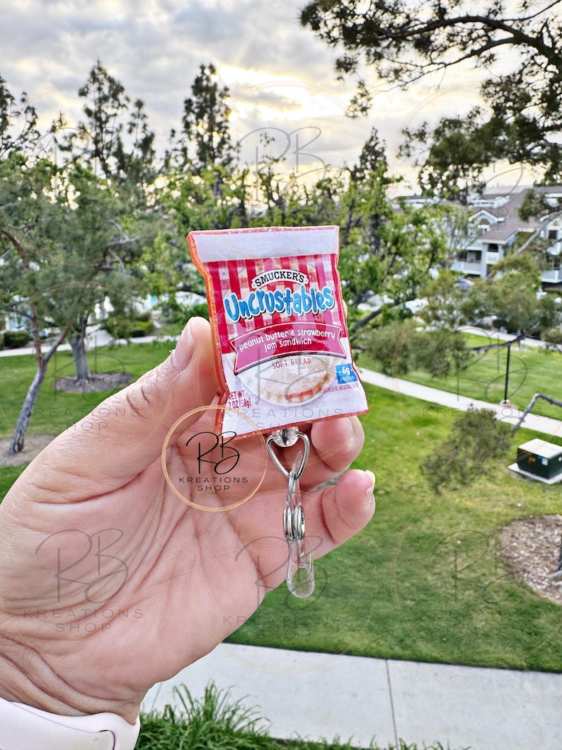 May include: A red and white Uncrustables peanut butter and strawberry jam sandwich keychain. The keychain features the Smucker's logo and the text "Uncrustables" in white letters. The keychain is attached to a silver metal clip.