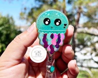 May include: A teal blue glitter resin jellyfish charm with iridescent purple and pink tentacles. The charm is attached to a clear plastic badge holder with a silver clip and a teal blue glitter resin bead.