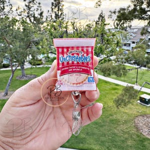 May include: A red and white Uncrustables peanut butter and strawberry jam sandwich keychain. The keychain is made to look like a real Uncrustables package.