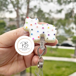 May include: A white, dog-shaped badge reel with colorful sprinkles and a gold glitter border. The reel has a clear plastic strap and a metal clip. The logo for RB Kreations Shop is visible.