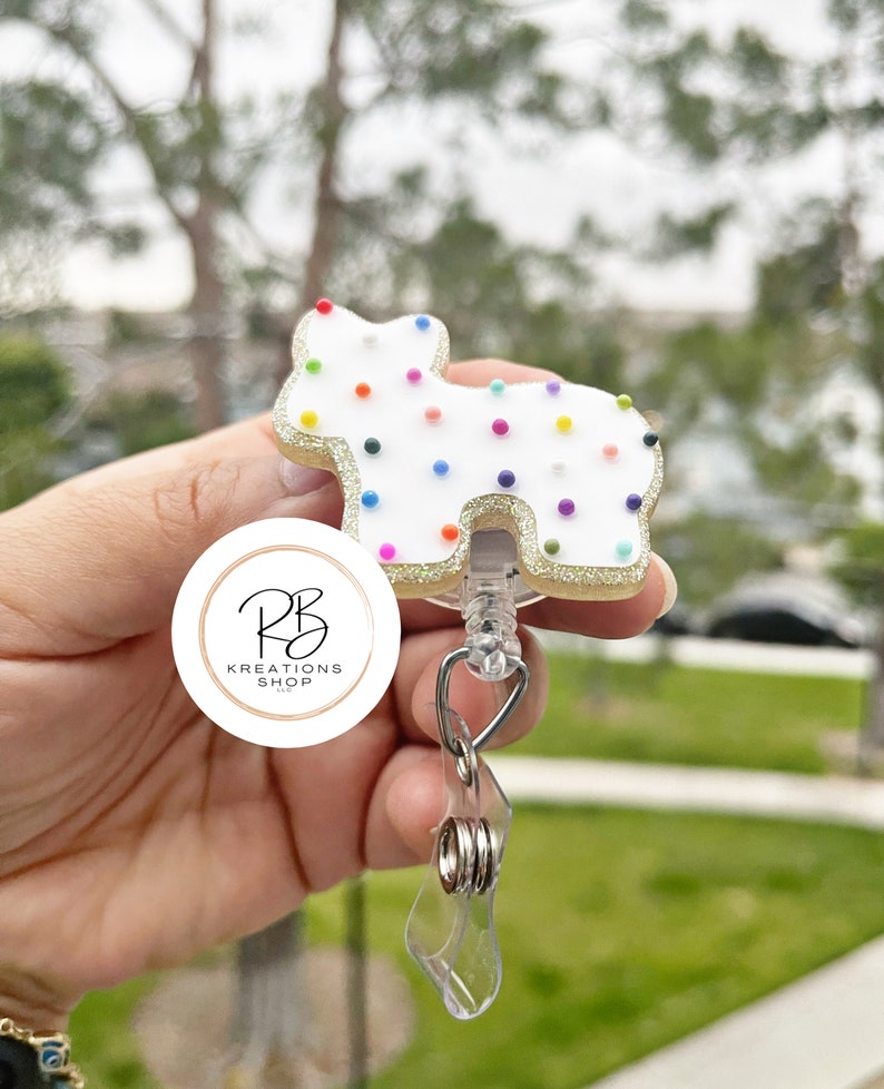 May include: A white, dog-shaped badge reel with colorful sprinkles and a gold glitter border. The reel has a clear plastic strap and a metal clip. The logo for RB Kreations Shop is visible.