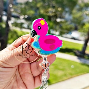 May include: A pink flamingo badge reel with a black beak and yellow details. The badge reel has a clear plastic clip and a retractable cord. The text "RB KREATIONS SHOP LLC" is visible on the badge reel.
