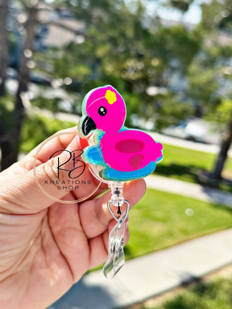 May include: A pink flamingo badge reel with a black beak and yellow details. The badge reel has a clear plastic clip and a retractable cord. The text "RB KREATIONS SHOP LLC" is visible on the badge reel.