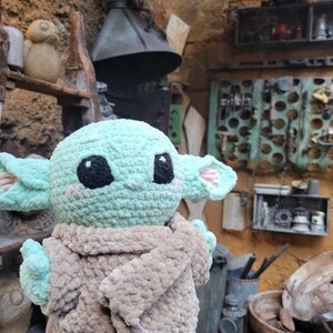 Puede incluir: Un juguete de peluche verde tejido a crochet con una manta de crochet marr&oacute;n. El juguete tiene ojos grandes y una boca peque&ntilde;a. El juguete est&aacute; sentado en la mano de una persona.