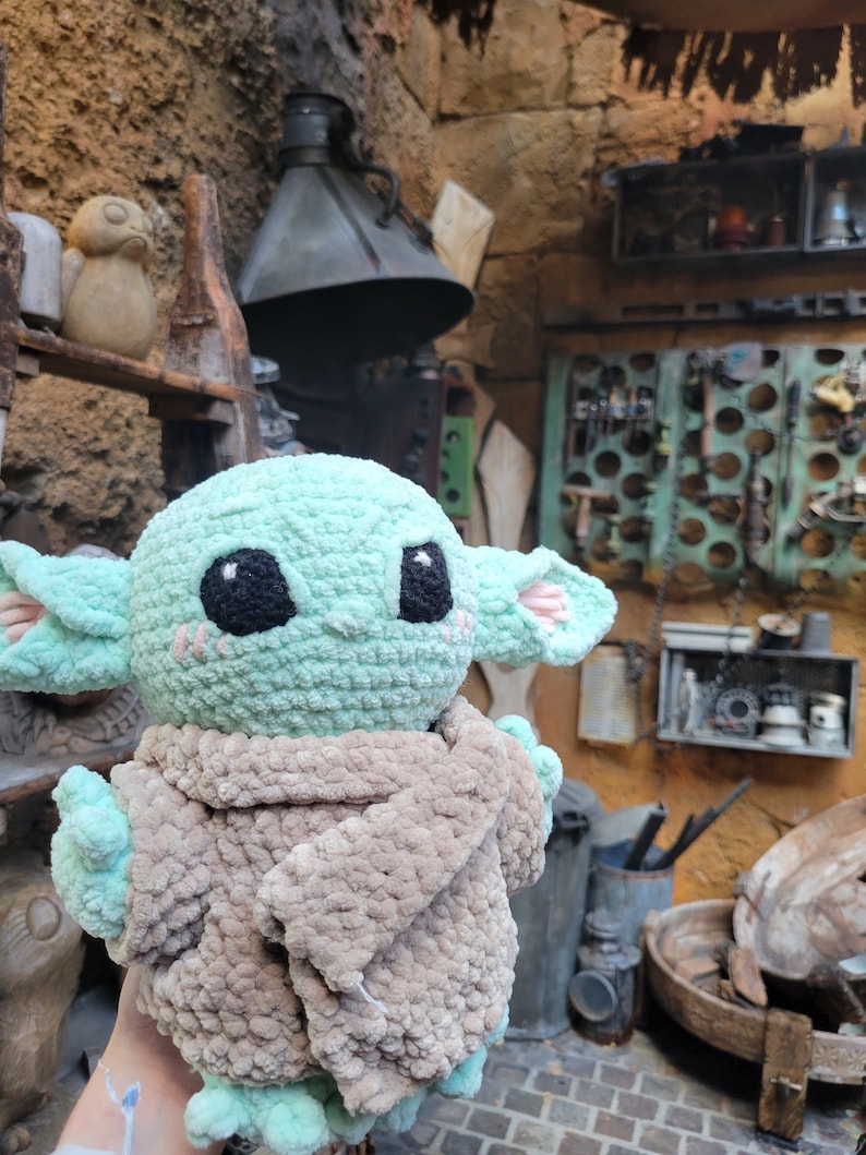 Puede incluir: Un juguete de peluche verde tejido a crochet con una manta de crochet marr&oacute;n. El juguete tiene ojos grandes y una boca peque&ntilde;a. El juguete est&aacute; sentado en la mano de una persona.