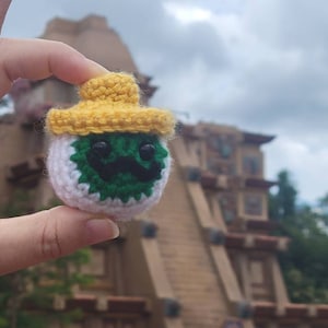 May include: A small crocheted figure wearing a yellow sombrero. The figure has a white face, green eyes, and a black mustache. The figure is being held in a person's hand.
