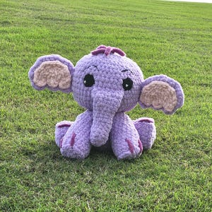 May include: A crocheted purple elephant plush toy with pink ears and a pink nose. The toy is sitting on a green grassy lawn.