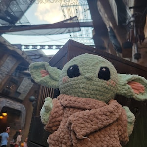 Puede incluir: Un juguete de peluche verde tejido a crochet de un personaje de la franquicia Star Wars, que lleva un abrigo tejido a crochet marr&oacute;n. El juguete est&aacute; sentado sobre una superficie de madera.