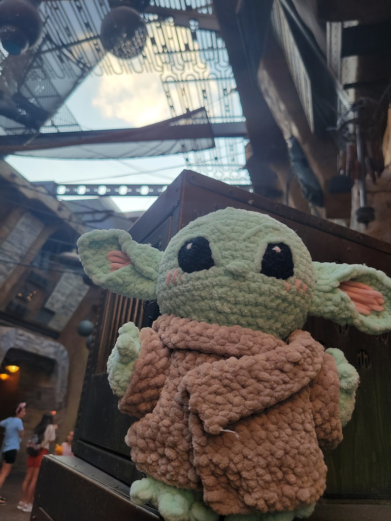 Puede incluir: Un juguete de peluche verde tejido a crochet de un personaje de la franquicia Star Wars, que lleva un abrigo tejido a crochet marr&oacute;n. El juguete est&aacute; sentado sobre una superficie de madera.