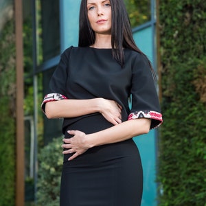 May include: Black two-piece outfit featuring a top and skirt. The top has short sleeves with patterned cuffs in red, white, and black. The skirt has a matching patterned detail at the hem. The outfit is a modern take on traditional design.