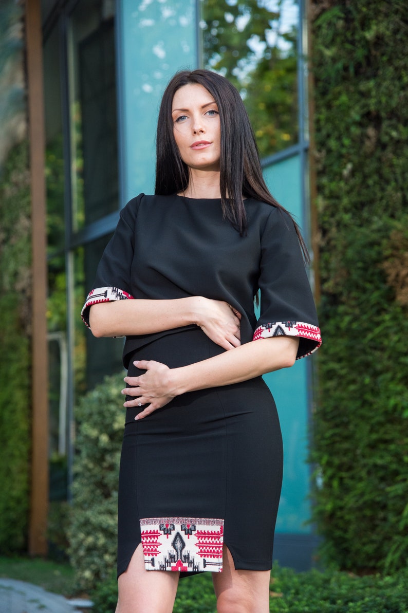 May include: Black two-piece outfit featuring a top and skirt. The top has short sleeves with patterned cuffs in red, white, and black. The skirt has a matching patterned detail at the hem. The outfit is a modern take on traditional design.