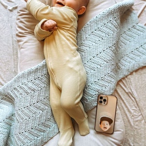 Op de afbeelding: Een lachende baby in een lichtgele geribbelde voetjesromper ligt op een wit kussen, bedekt met een lichtblauwe gehaakte deken. Een telefoon met een aangepaste pop-up grip met een kindergezicht ligt in de buurt.