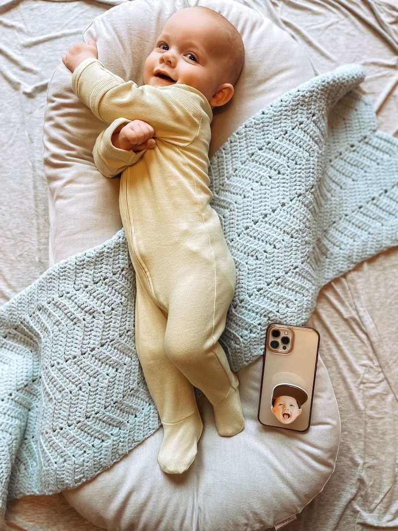 Op de afbeelding: Een lachende baby in een lichtgele geribbelde voetjesromper ligt op een wit kussen, bedekt met een lichtblauwe gehaakte deken. Een telefoon met een aangepaste pop-up grip met een kindergezicht ligt in de buurt.