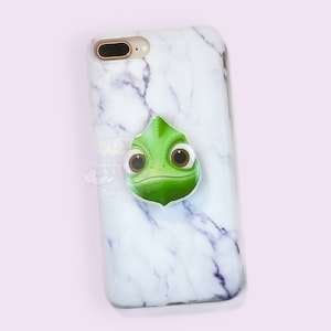 Puede incluir: Una funda de teléfono de mármol blanco con un soporte pop de camaleón verde. El camaleón tiene grandes ojos marrones y un cuerpo verde.