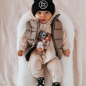 Op de afbeelding: Een baby met een zwarte muts met de tekst "HUG LIFE", een beige gewatteerde bodywarmer en een bijpassende jumpsuit. De baby draagt ook zwart-witte sneakers en houdt een telefoonhoesje met foto's vast.