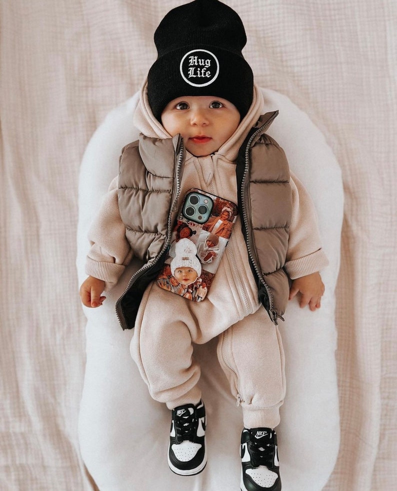 Op de afbeelding: Een baby met een zwarte muts met de tekst "HUG LIFE", een beige gewatteerde bodywarmer en een bijpassende jumpsuit. De baby draagt ook zwart-witte sneakers en houdt een telefoonhoesje met foto's vast.