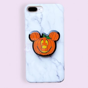 Puede incluir: Una funda de teléfono de mármol blanco con un agarre de teléfono de calabaza Mickey Mouse negro. La calabaza es naranja con un tallo verde y un contorno negro.