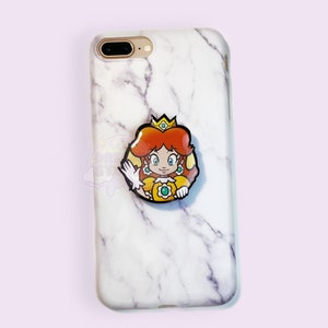 Puede incluir: Una funda de teléfono de mármol blanco con un soporte pop que presenta a la Princesa Peach de la serie de videojuegos Super Mario Bros. El soporte pop es amarillo, naranja y verde.