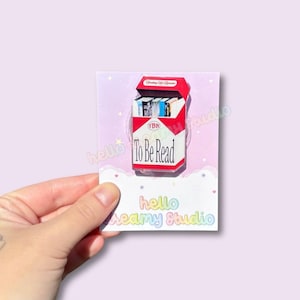 May include: A rectangular enamel pin shaped like a cigarette pack with a red and white design. The pack features the text To Be Read and contains a row of miniature books instead of cigarettes. The pin is attached to a light purple backing card.
