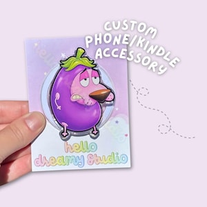 May include: A purple eggplant-shaped character accessory with a green stem, large tired eyes, and a brown nose. The backing card features the text CUSTOM PHONE/KINDLE ACCESSORY and hello dreamy studio in pastel rainbow lettering on a light purple background.