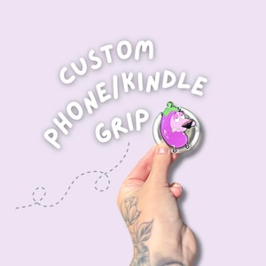 May include: A custom phone/Kindle grip featuring a cartoon eggplant character with a worried expression. The grip is round with a clear border and is held against a light purple background. The text "CUSTOM PHONE/KINDLE GRIP" is in white.
