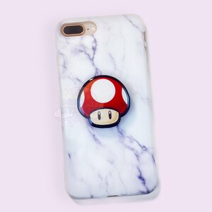 May include: White marble iPhone case with a red and white mushroom pop socket.