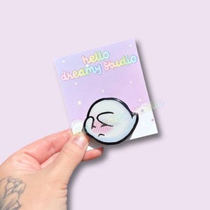 Puede incluir: Una tarjeta de color pastel con un fantasma de dibujos animados blanco con contorno negro y expresión triste. La tarjeta tiene el texto "hello dreamy Studio" en arcoíris. El fondo es morado suave con pequeñas estrellas.